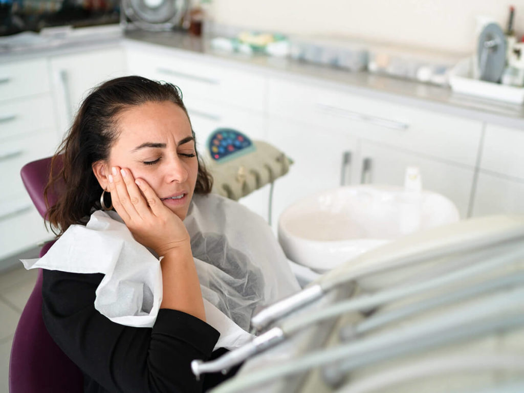 A patient in dental distress, clutching her cheek and wincing in pain, sits in a dental chair, indicating a potential emergency dental situation, in a clinic setting.