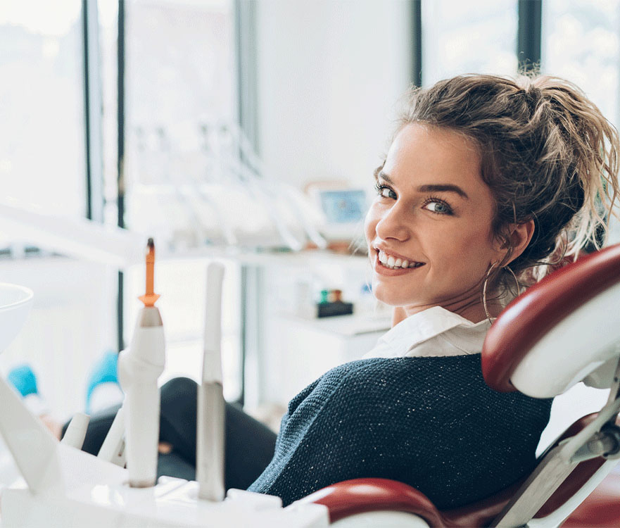 Portrait of a cheerful young woman with a ponytail sitting in a dental chair, smiling brightly, showcasing her healthy teeth, with dental equipment visible in the background, indicating a positive dental visit experience."