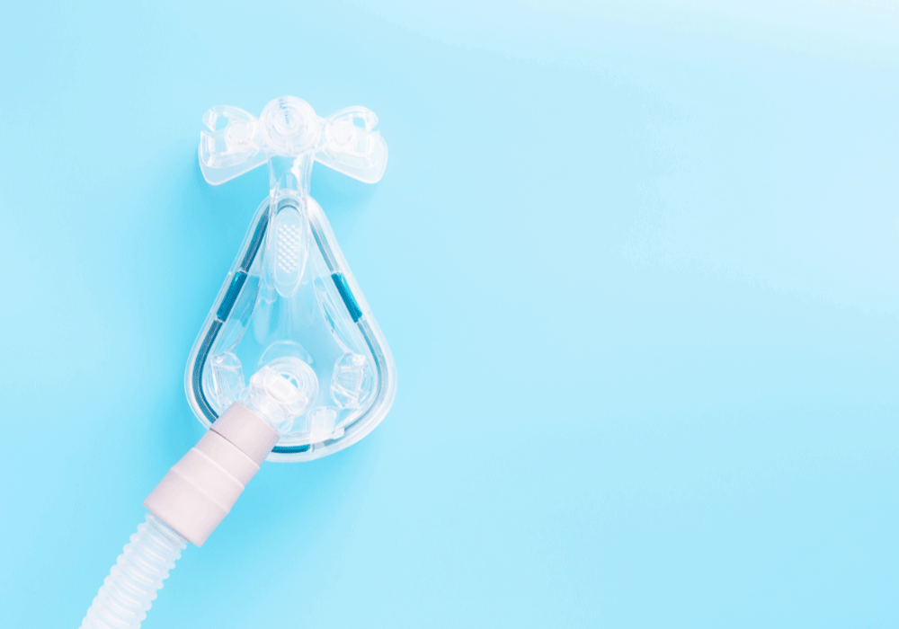 Close-up image of a transparent sleep apnea mask placed on a light blue background, highlighting the mask's detailed structure with tubing and adjustment straps, emphasizing medical respiratory assistance.